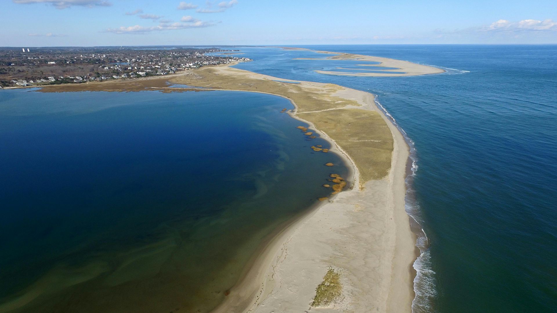 Aerial view of a narrow, sandy barrier beach separating a calm, dark blue bay from the open, deeper blue ocean.