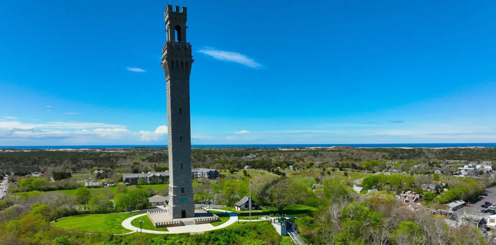 A tall, granite Pilgrim Monument stands on a green hilltop under a clear blue sky, overlooking a coastal landscape.