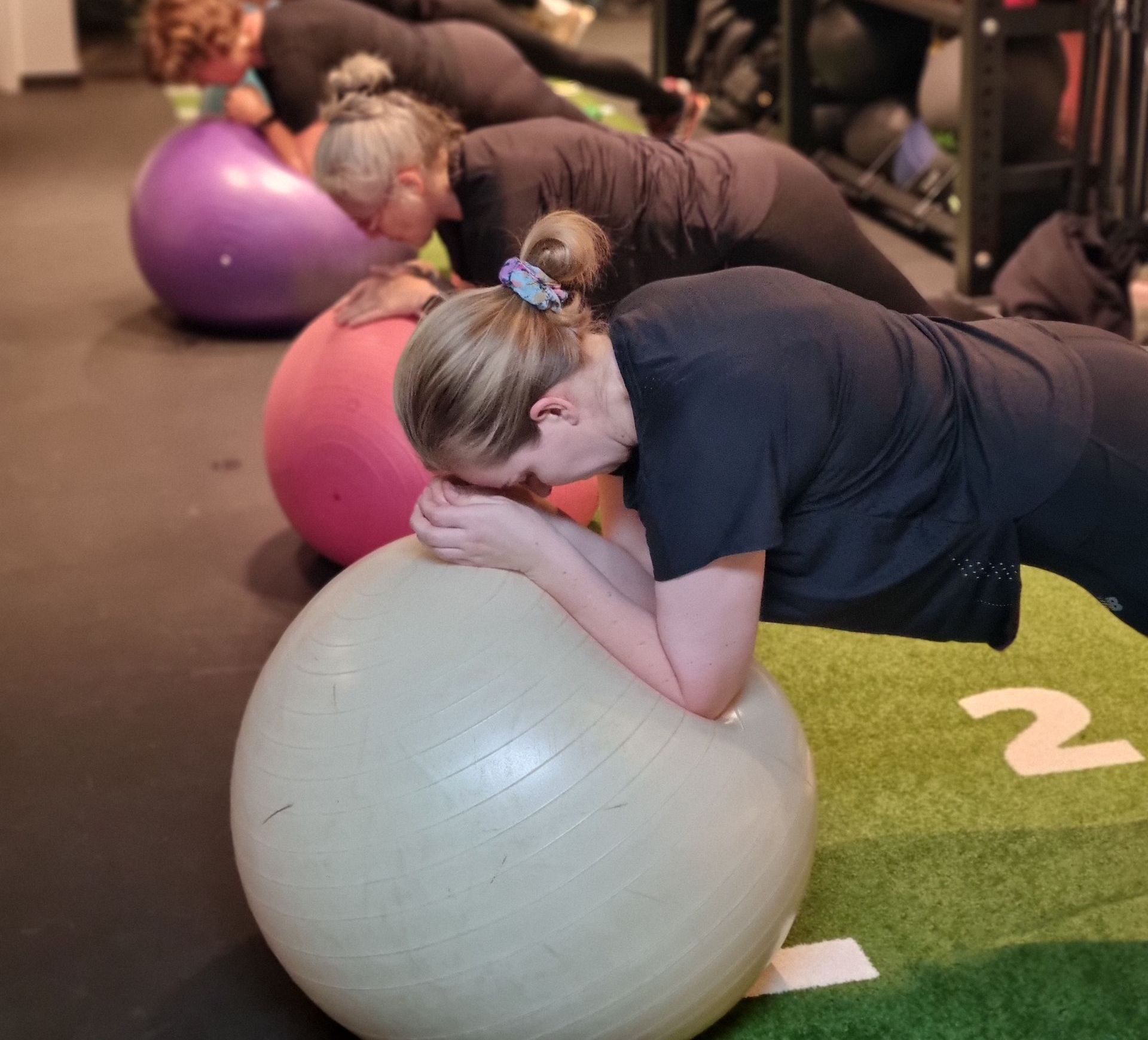Mensen die plankoefeningen doen op fitnessballen in een sportschool, met groen kunstgras en genummerde vloermarkeringen.