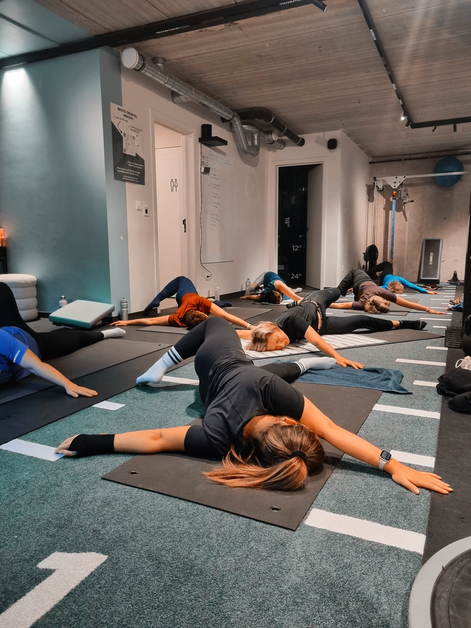 Mensen doen rek- en strekoefeningen op yogamatten in een fitnessstudio met een grijze vloer en witte muren.