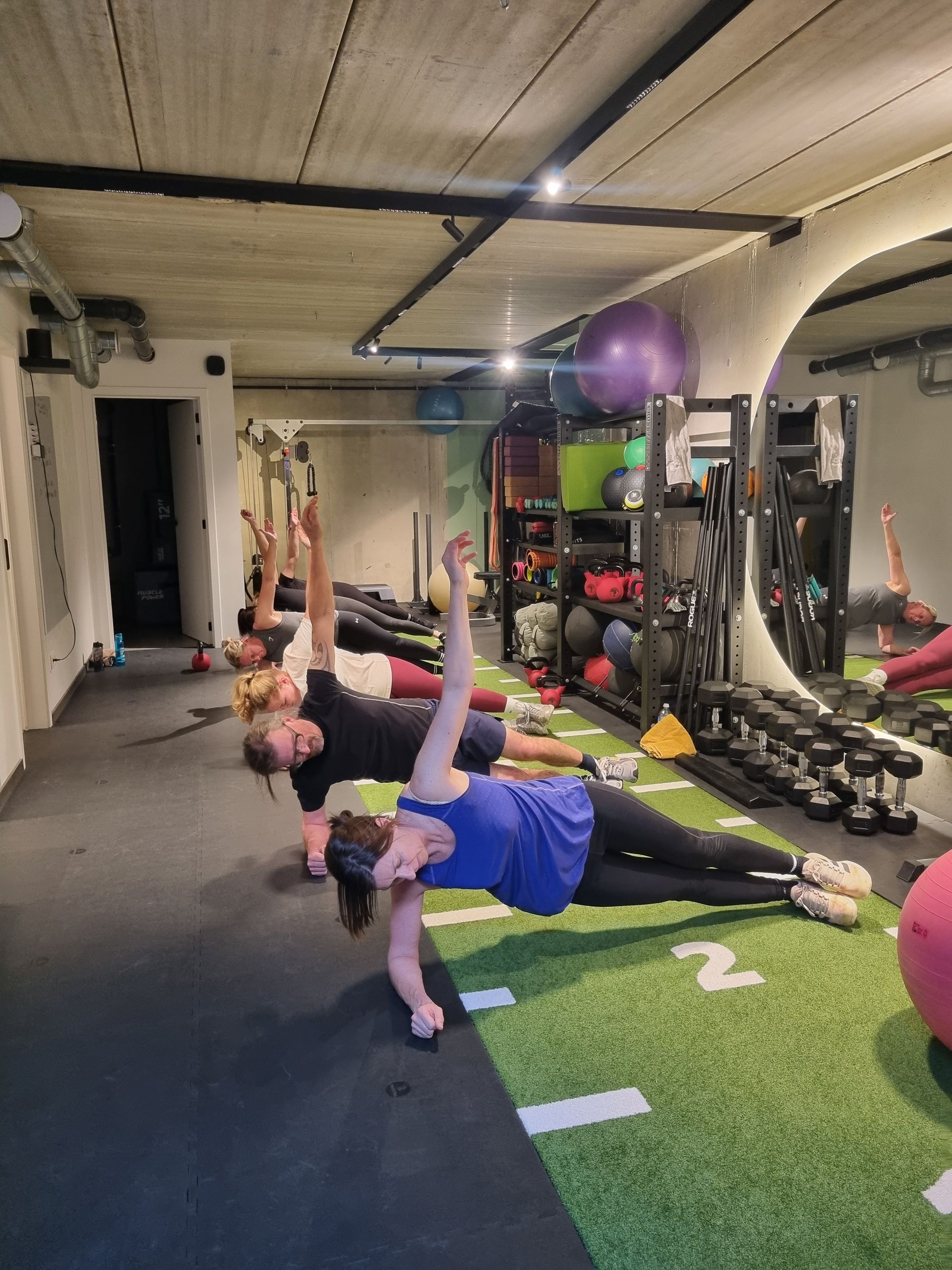 Mensen doen vloeroefeningen in een sportschool met kunstgras, dumbbells en fitnessballen.