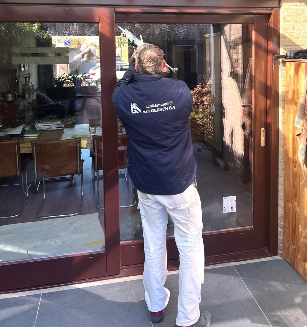 Man in blauw jasje en witte broek werkt aan een glazen schuifdeur buiten.