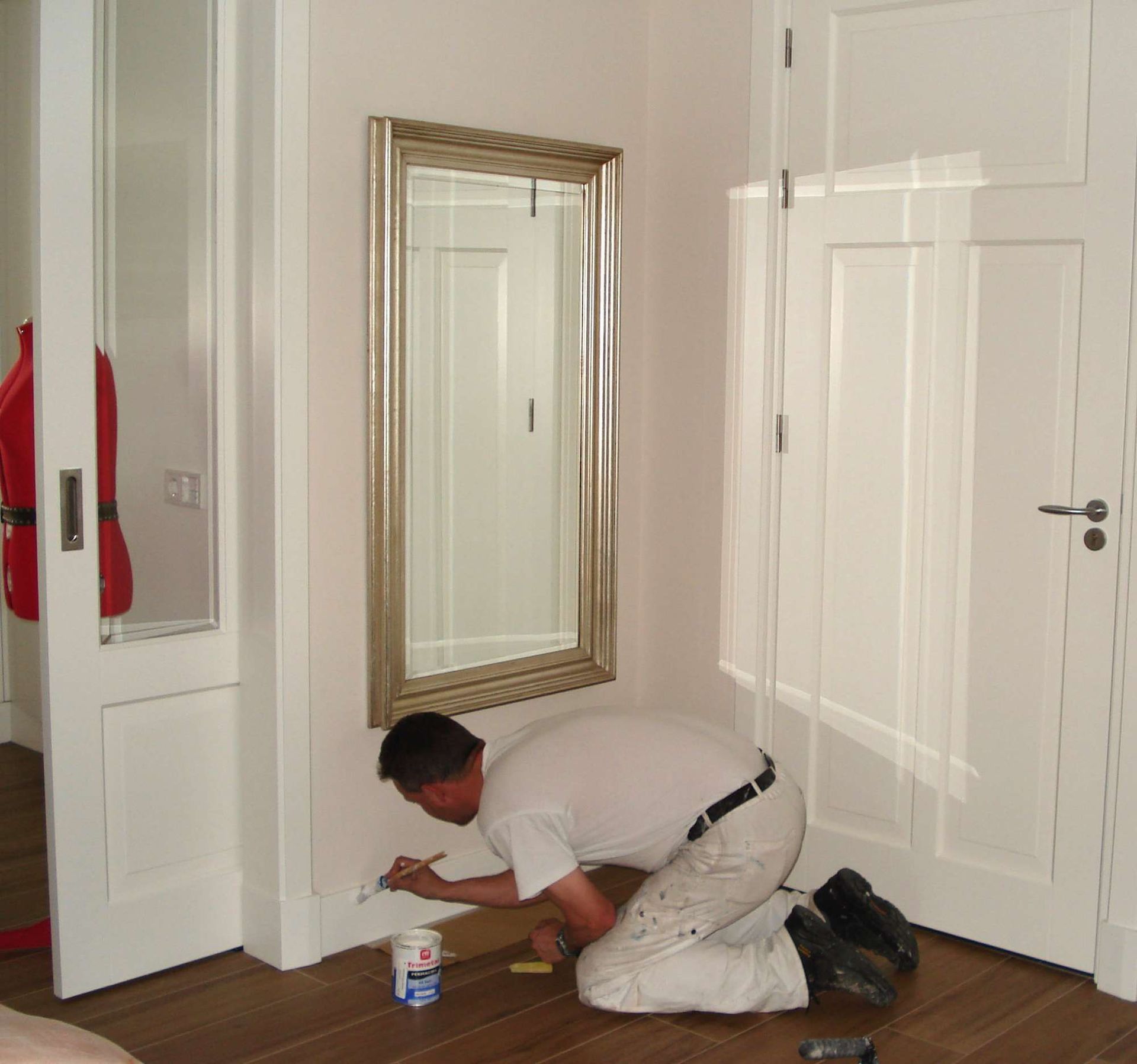 Een man schildert een witte plint in een kamer met een spiegel en deuren.