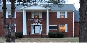 Brick county office building with white-trimmed entrance and lit windows, framed by two trees