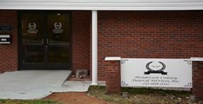 Entrance to a brick office building with glass doors and a white sign by the walkway.