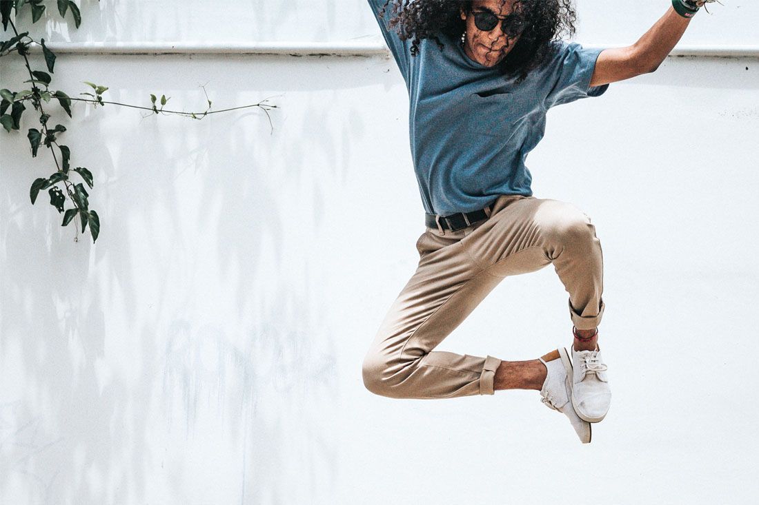 A person with curly hair and sunglasses jumps mid-air against a white wall with leafy green vines on the left.