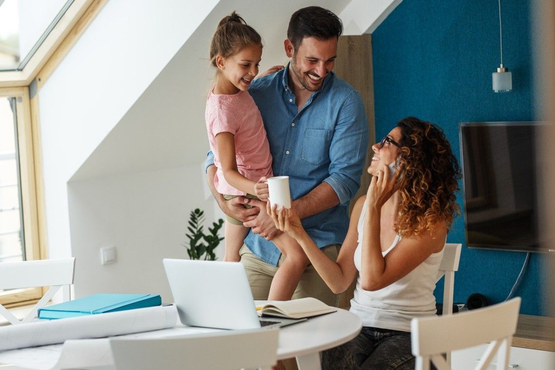 A family gathers in a bright room, where an adult on a call sits by a laptop while another holds a child near a table.
