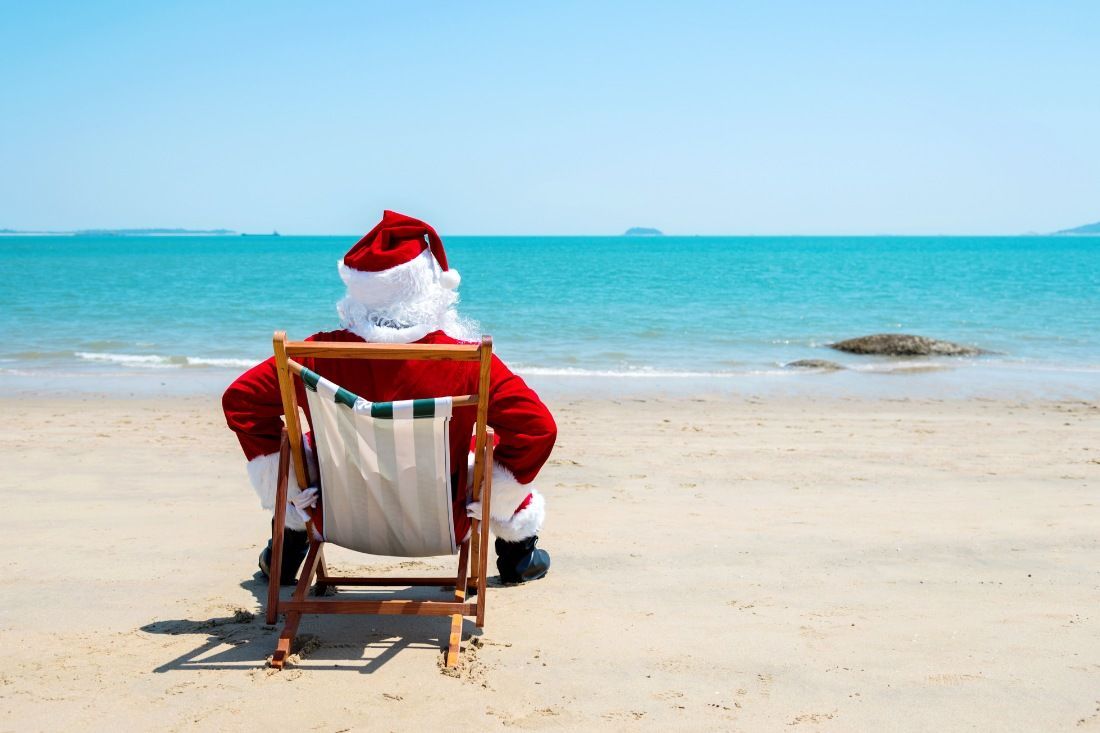 Santa Claus sits in a beach chair on the sand, looking out at the turquoise ocean on a sunny day.