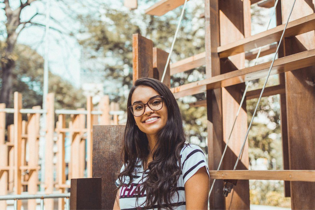 A smiling person with long dark hair and glasses wears a striped shirt, standing outdoors in front of a wooden structure.