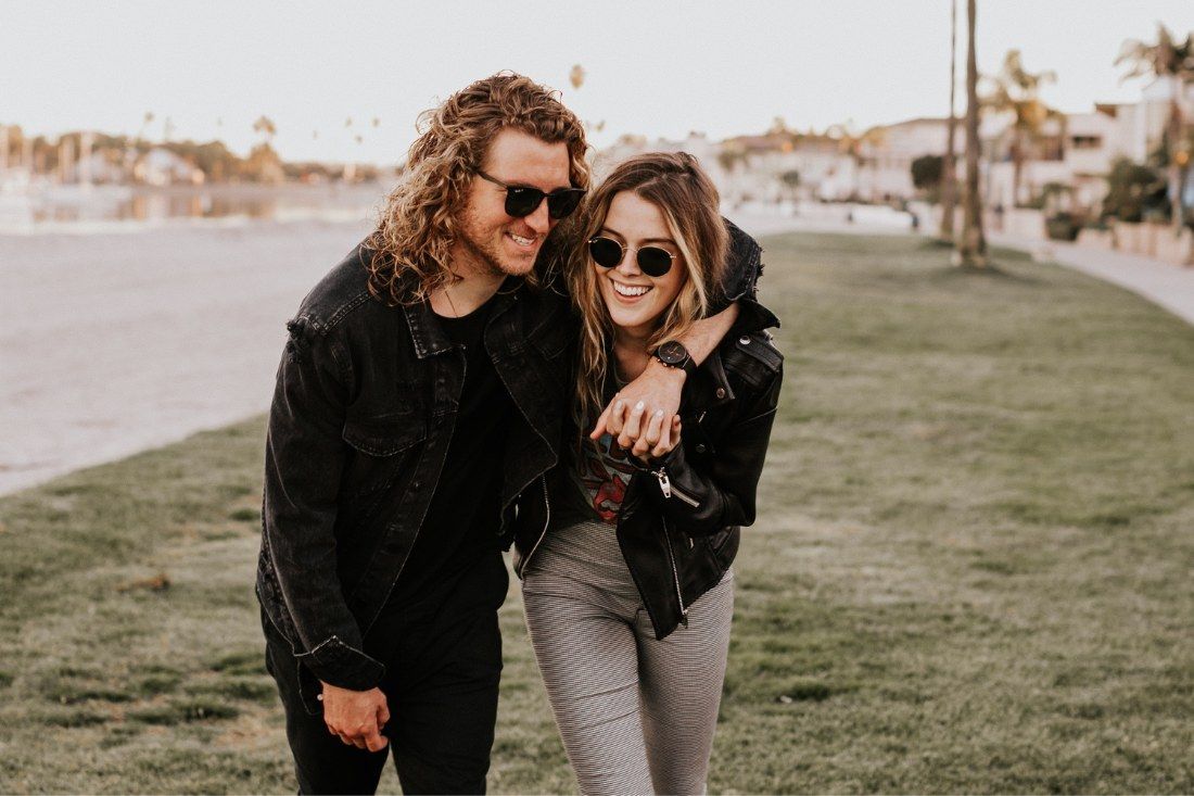 A smiling couple with long curly hair and sunglasses, wearing black jackets, walking together on a grassy area.
