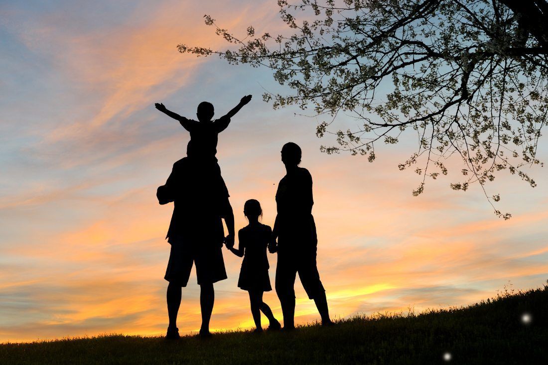 Silhouetted family of four standing on a hill at sunset, with a child held on shoulders, against a colorful sky.