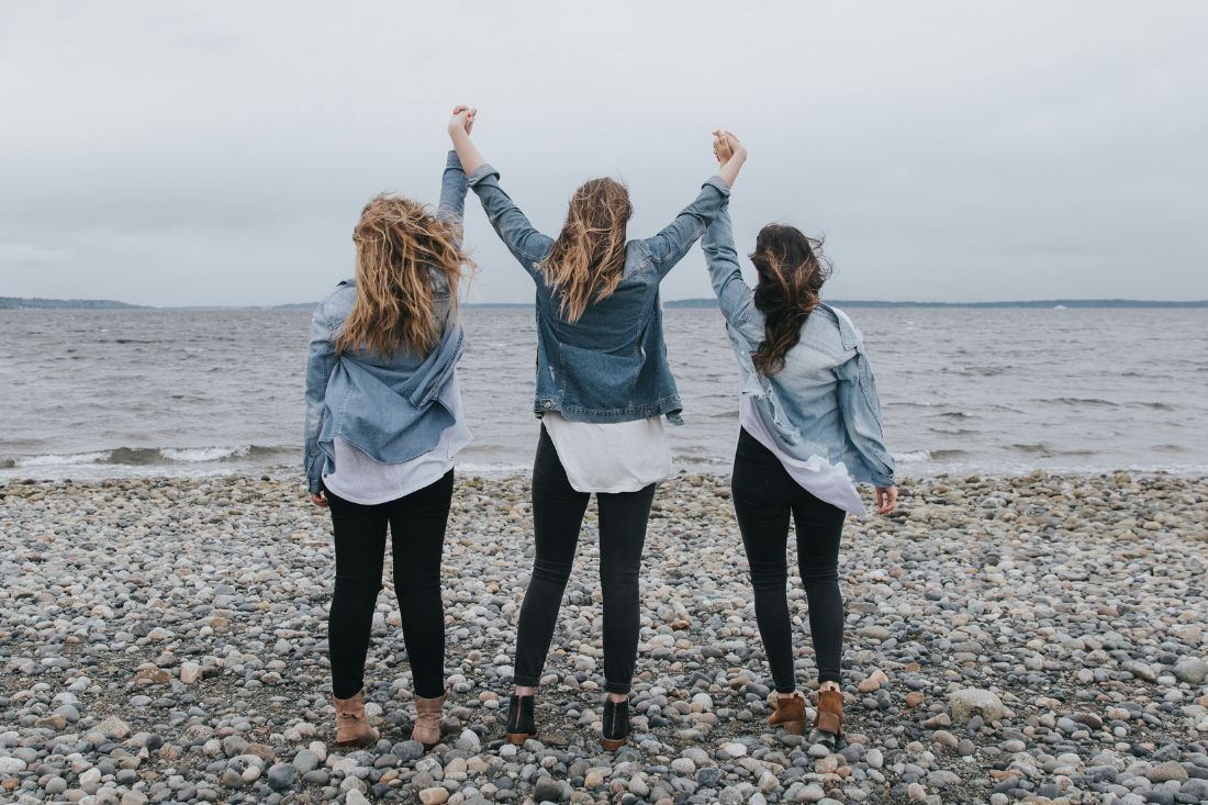 Three people with their arms raised stand in denim jackets and dark pants on a rocky beach facing the water.