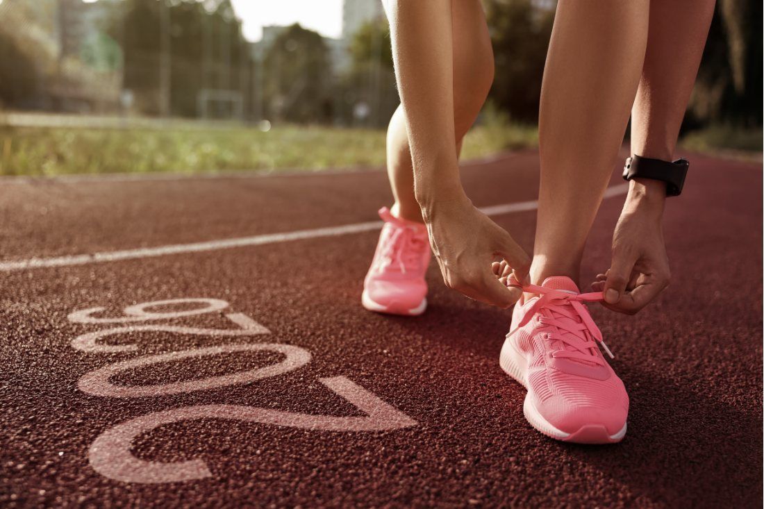 A runner ties their bright pink sneakers on a red track marked with the year 2026.