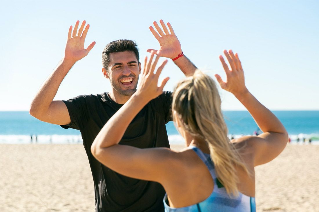 A man and woman face each other on a beach with hands raised to high-five, both smiling in the sunlight.