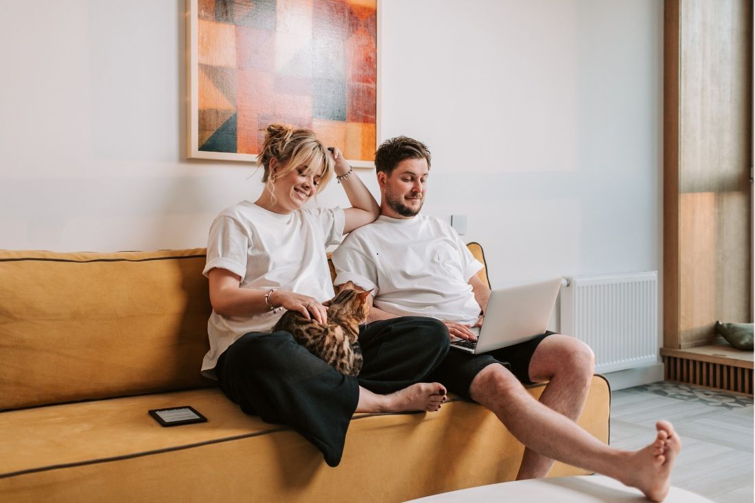A person sitting on a yellow sofa pets a cat while another person beside them uses a laptop in a bright room.