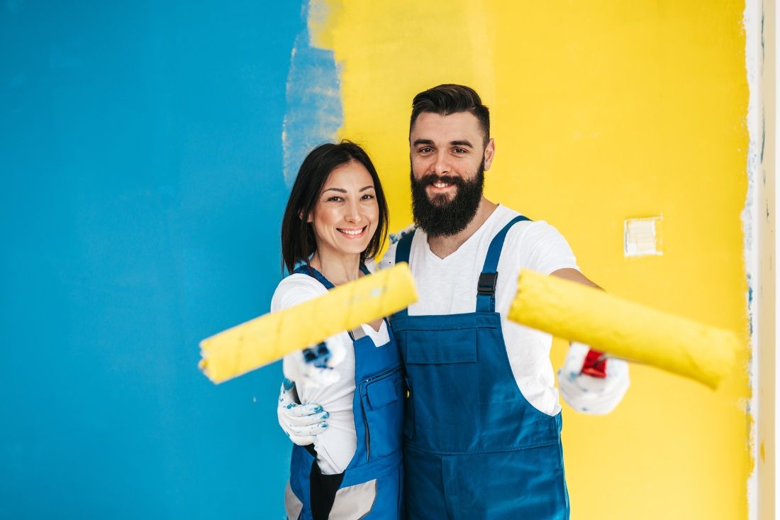 A smiling couple in blue overalls holding yellow paint rollers in front of a wall painted partially blue and yellow.