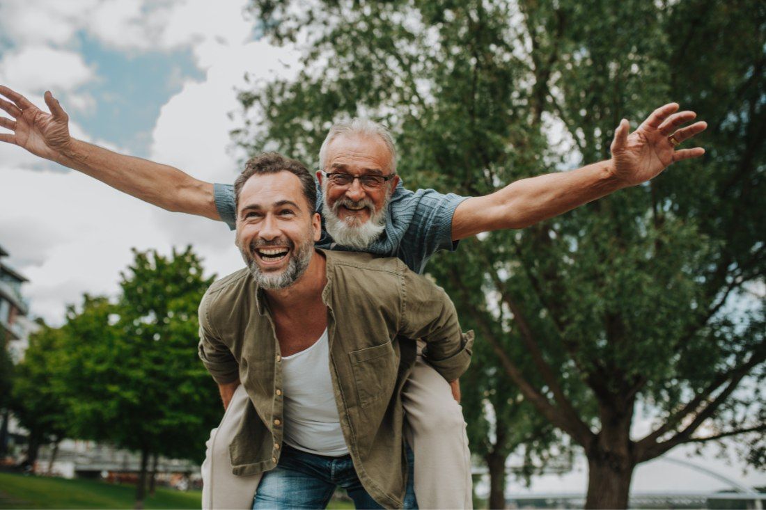 A man gives another man a piggyback ride outdoors with arms outstretched in a joyful gesture against a backdrop of trees.