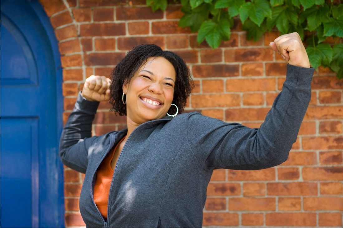 A smiling person in a gray jacket poses flexed in front of a brick wall and a blue door.