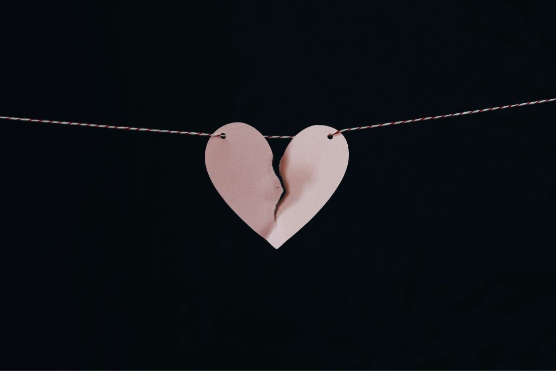 A light pink, heart-shaped paper cutout, torn in two, hangs from a thin string against a solid black background.