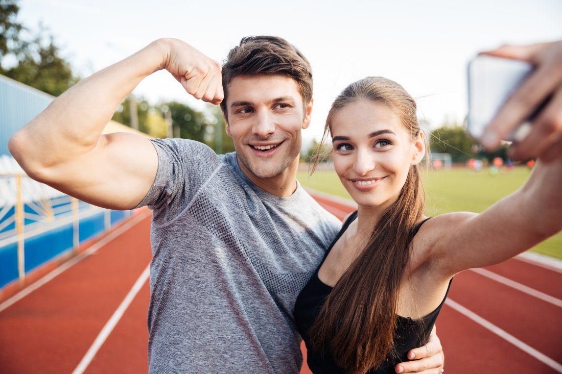Two people smile for a selfie on an outdoor track, with one person flexing their arm muscle.
