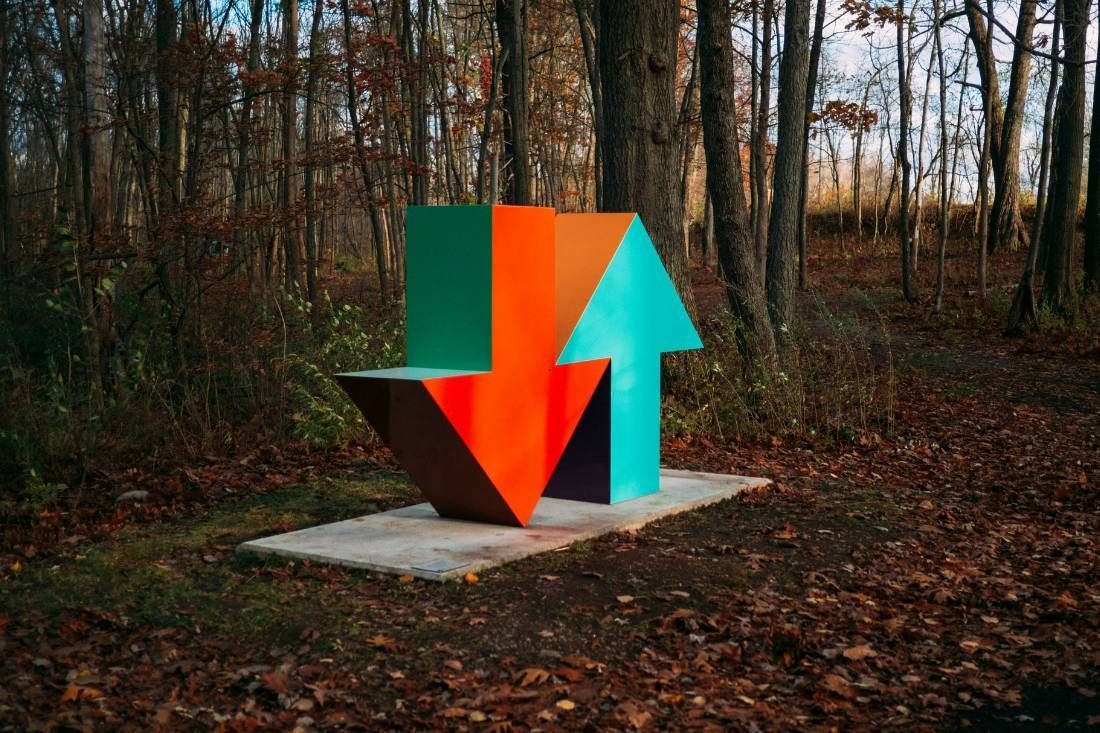 A large, abstract sculpture of a red downward arrow and a teal upward arrow standing on a concrete pad in a wooded area.