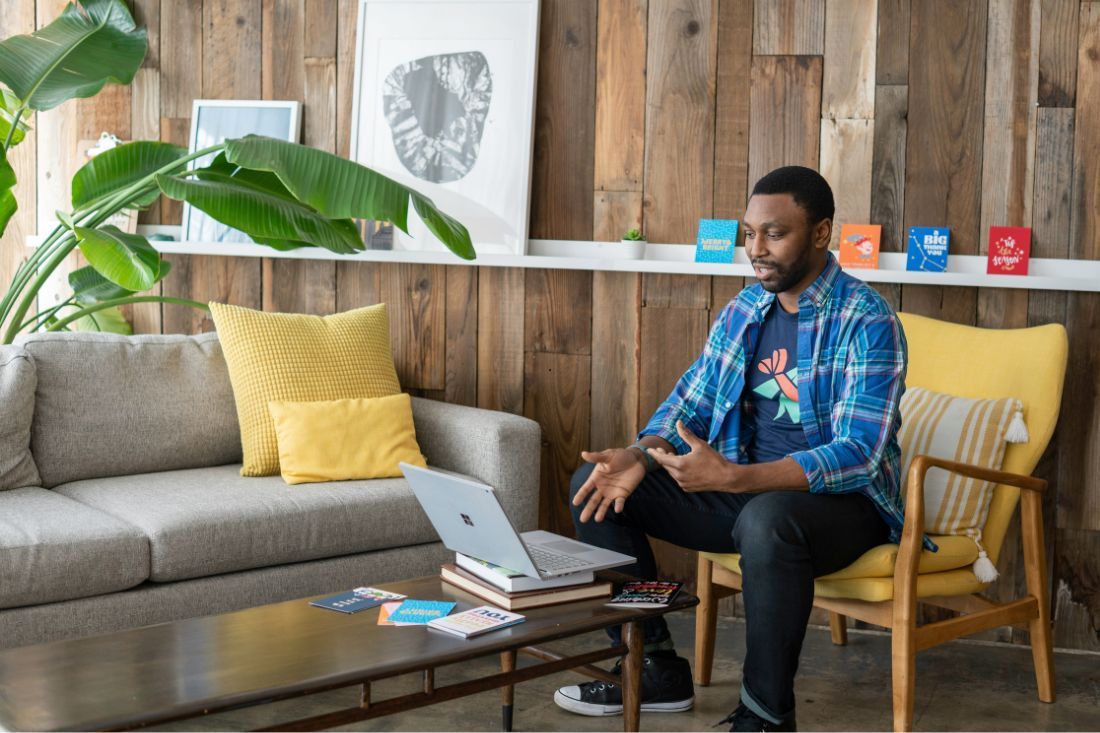 A person sitting in a yellow chair, talking during a video call on a laptop in a rustic, wood-paneled living room.
