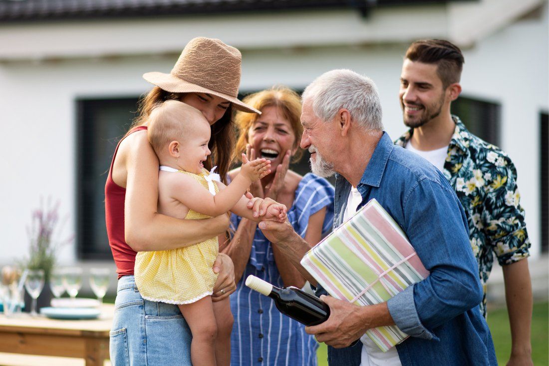 Family members gathered outdoors, smiling and interacting with a baby, with an adult holding a gift and a wine bottle.