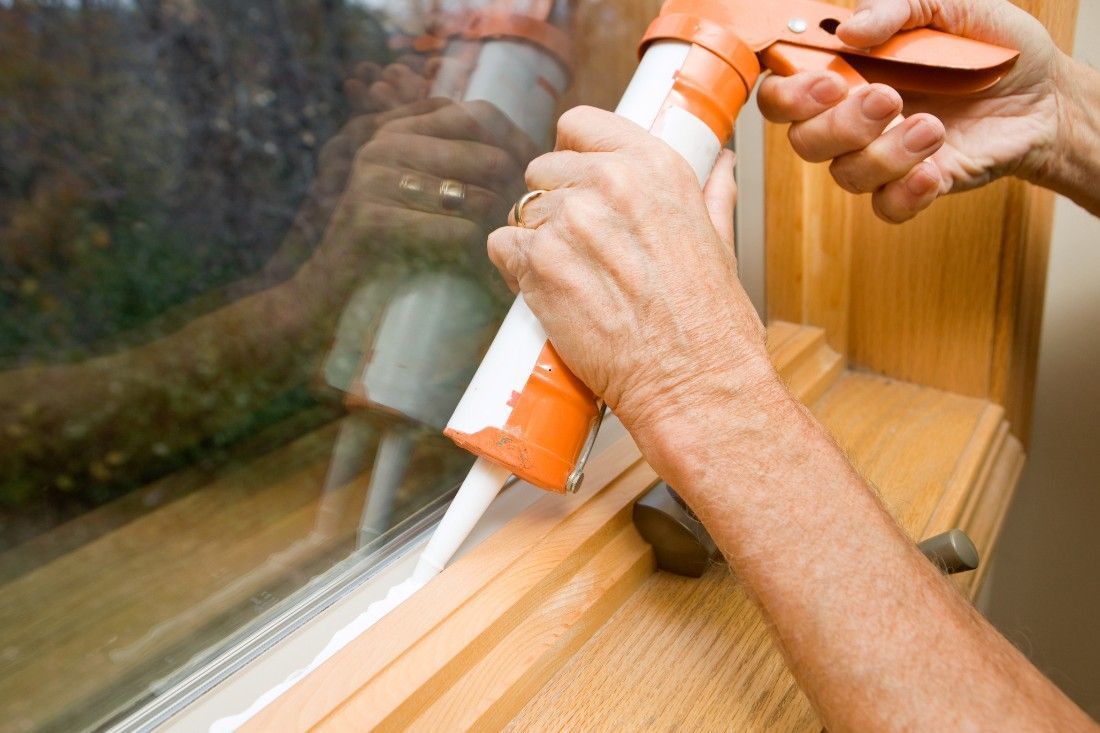 A person applying white caulk from a handheld orange caulk gun along the edge of a wooden window frame.