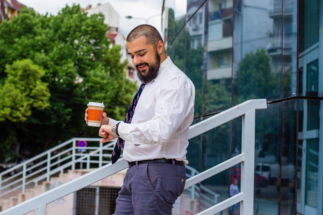 A bearded person in a white shirt and tie stands on outdoor stairs, holding a coffee cup and checking their watch.
