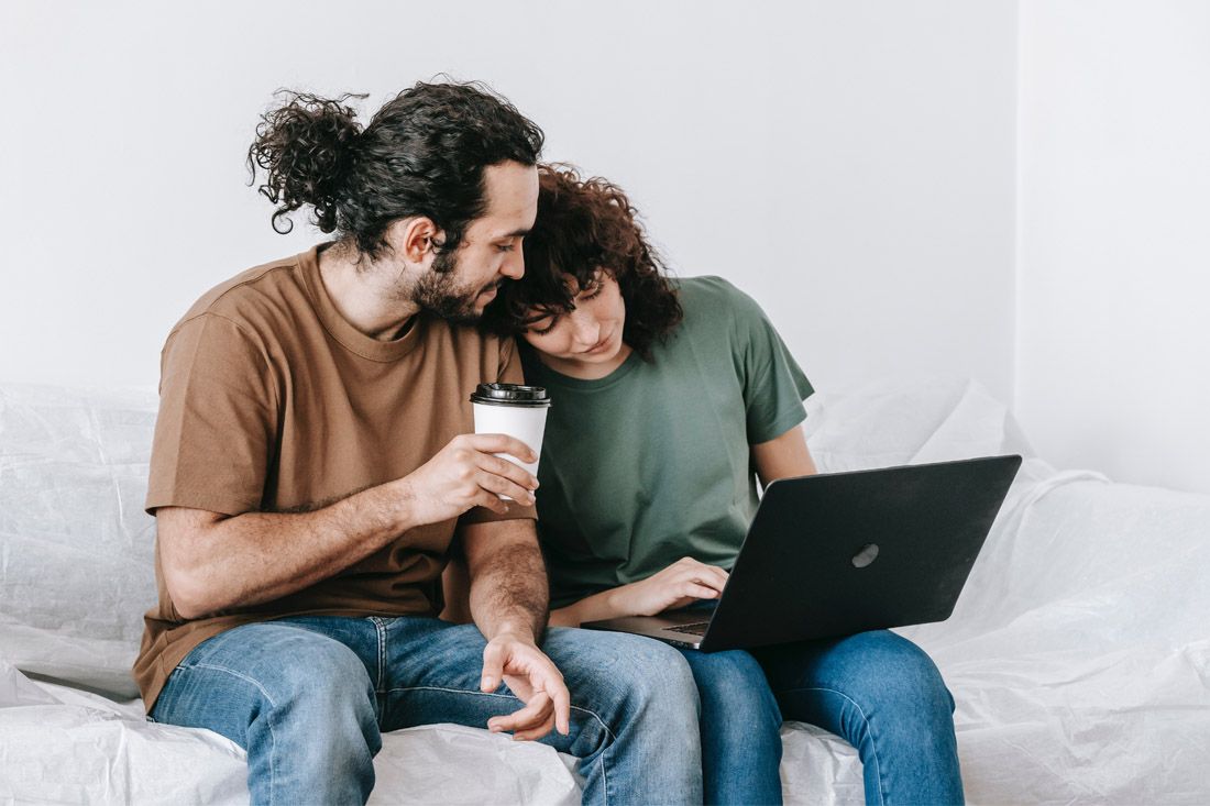 Two people sit together on a sofa, looking at a laptop while one holds a coffee cup and the other types.
