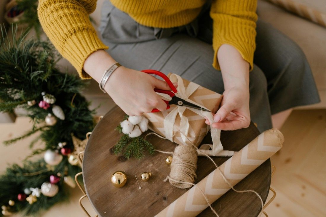 A person in a yellow sweater wraps a gift in brown paper with ribbon on a wooden table beside a decorated wreath.