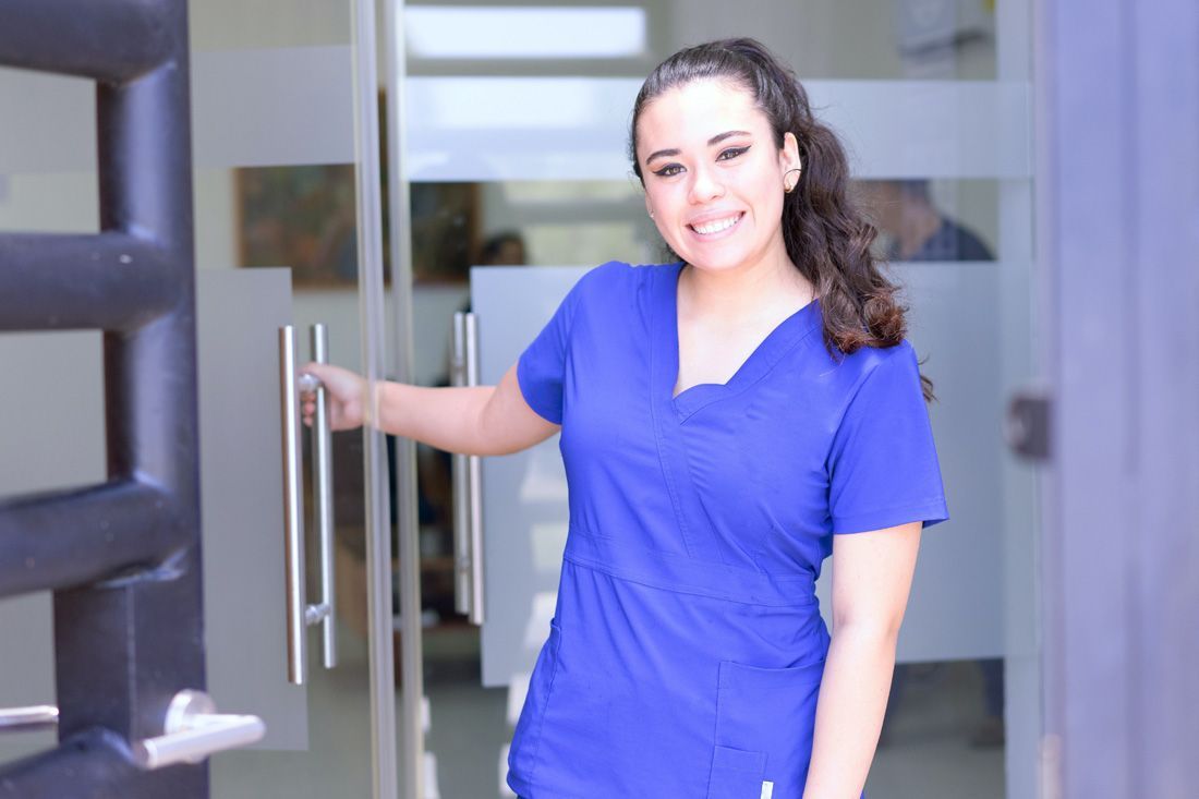 A smiling person in a blue scrub top holding open a glass door to a building.