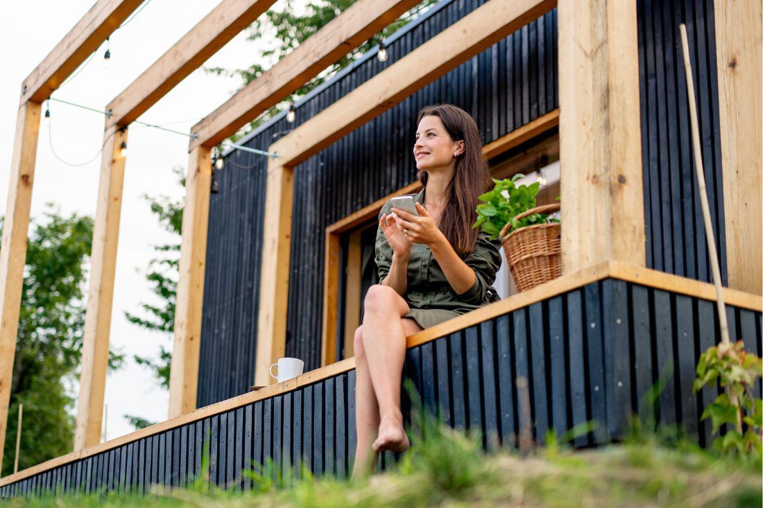 A person sits on the wooden porch of a modern cabin with dark siding, holding a phone and looking away with a smile.
