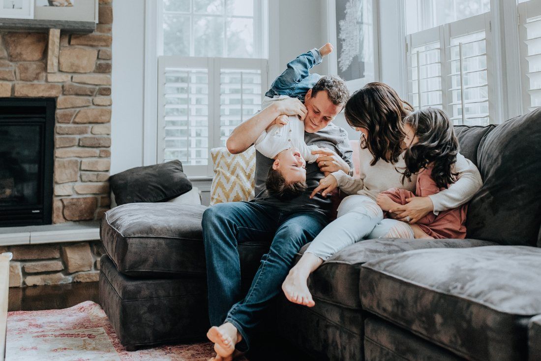 A family sits on a gray sofa in a living room, with a parent playfully holding a child upside down above them.