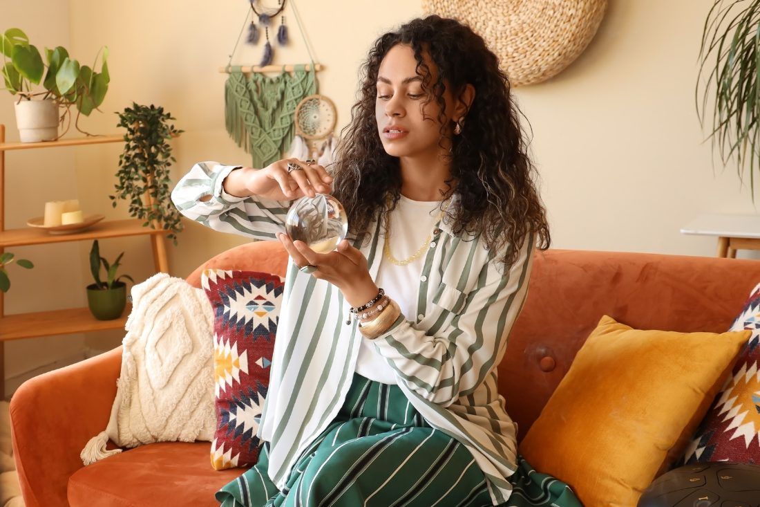 A person with curly hair sits on an orange sofa, holding a glass orb in a room decorated with plants and wall hangings.