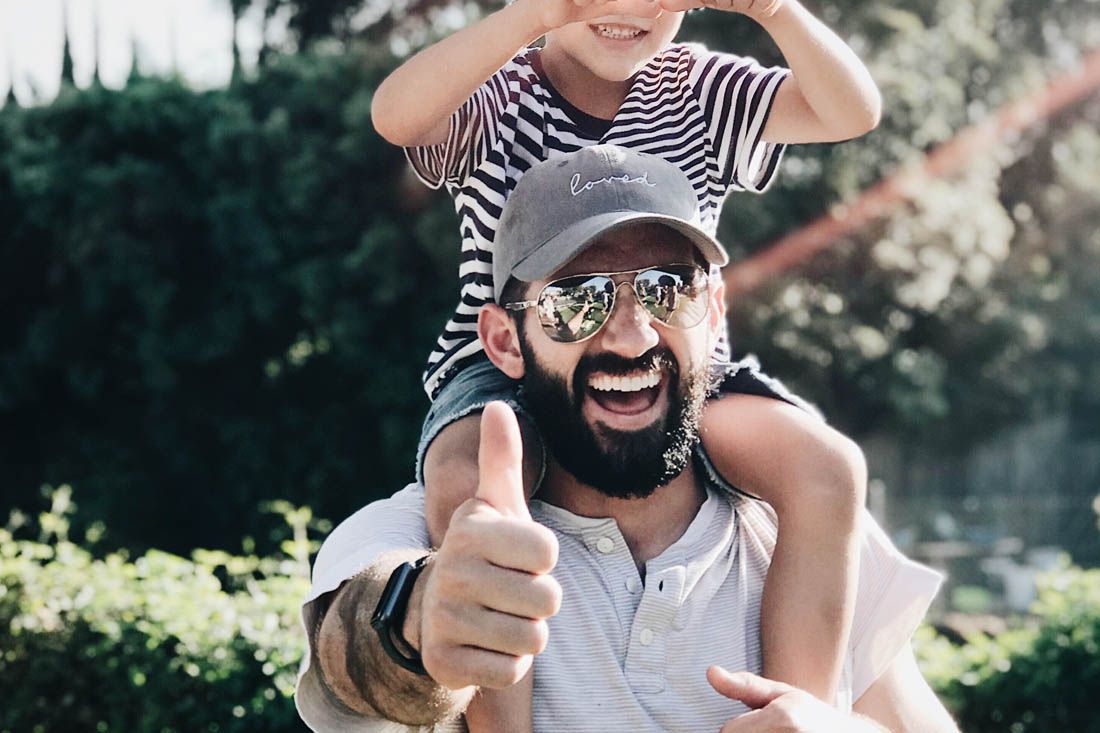 A bearded person wearing sunglasses and a cap gives a thumbs up while carrying a child on their shoulders outdoors.