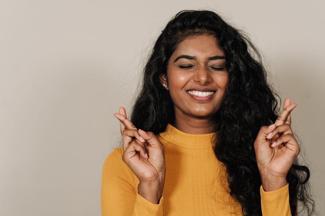 A smiling person with long dark wavy hair, wearing a mustard yellow top, holding both hands up with fingers crossed.