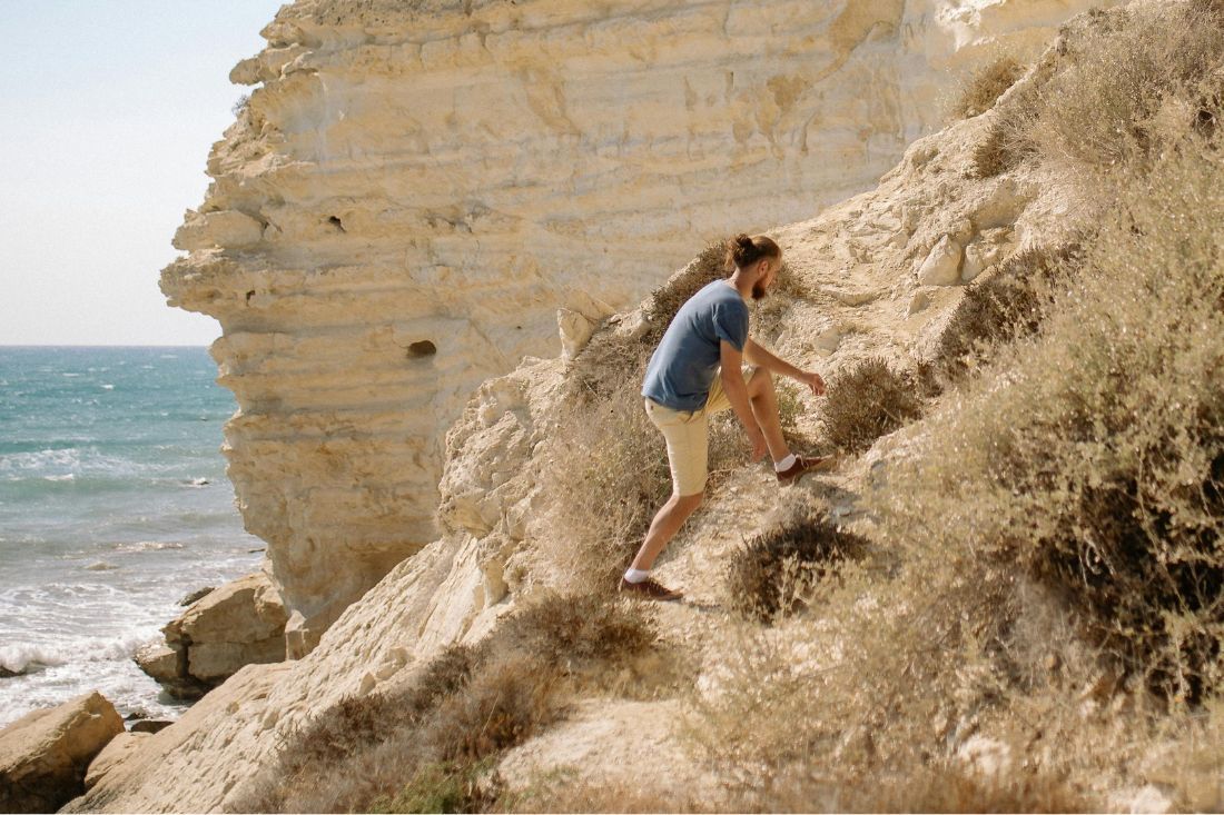 A person in a blue shirt and light shorts climbs a rugged, light-colored rocky cliffside overlooking the ocean.