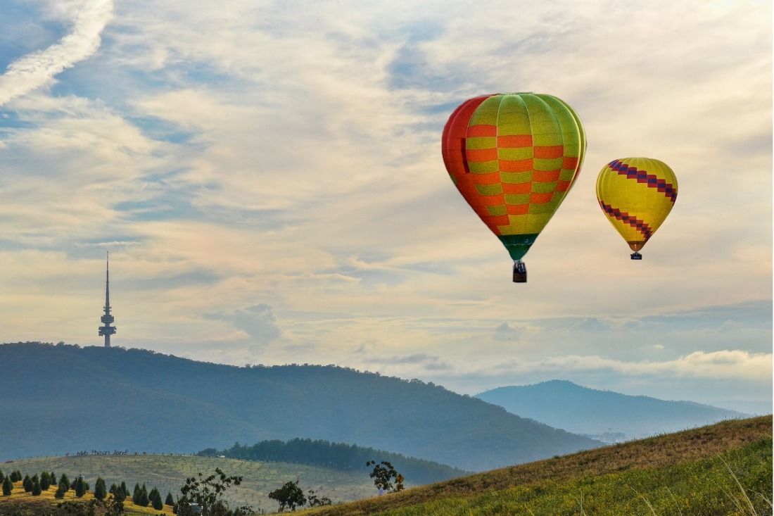 Two hot air balloons float over a grassy hill with a distant tower and mountains under a cloudy sky.