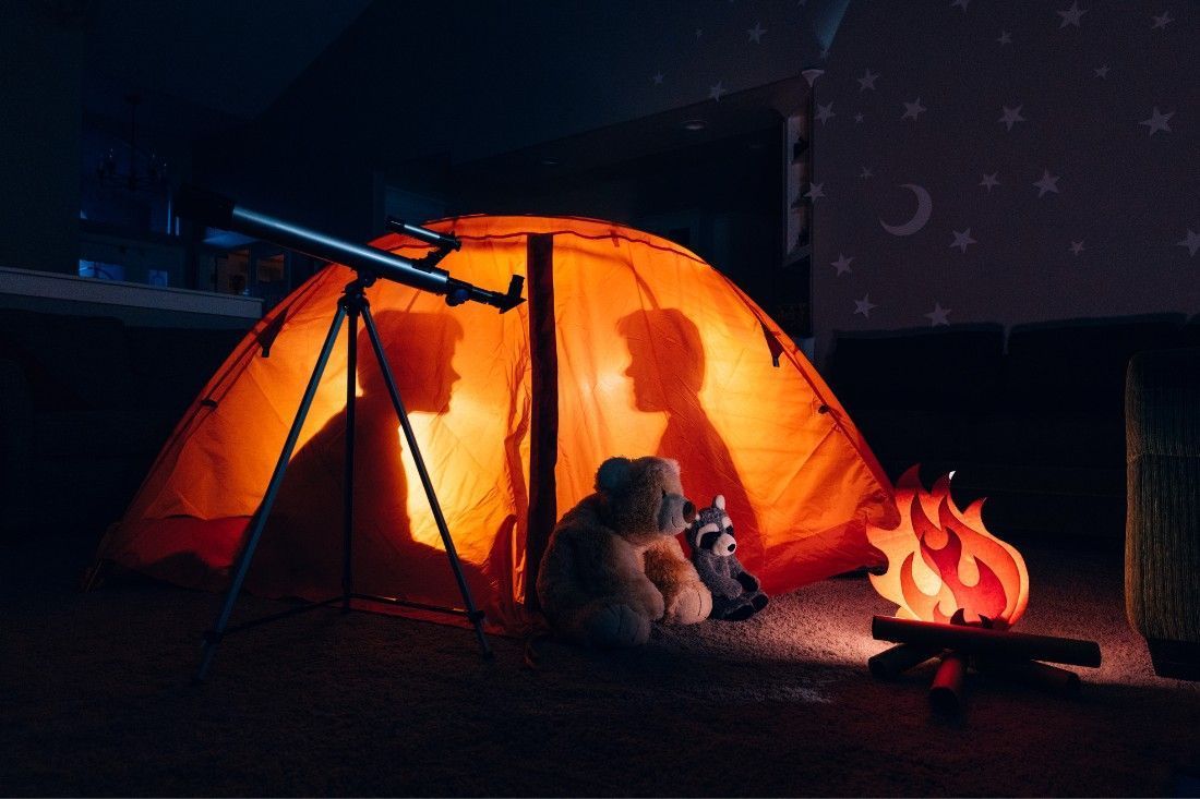 An orange indoor tent lit from within, showing two silhouettes, with a toy campfire and telescope in a dark room.
