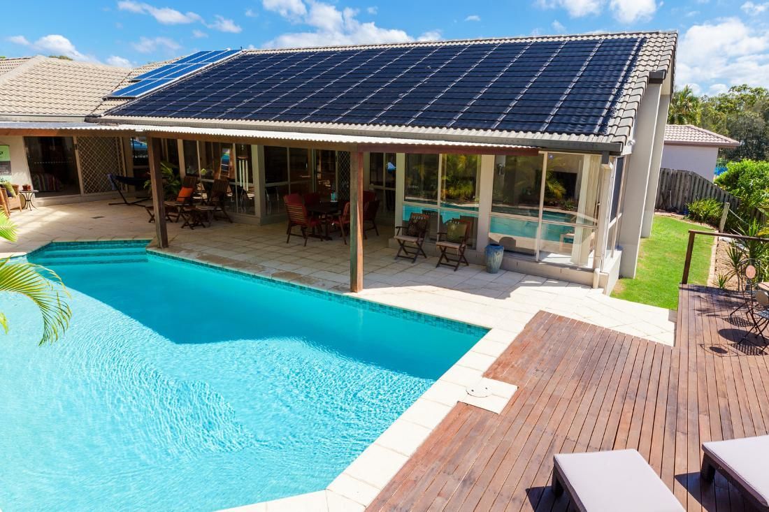 A bright blue swimming pool sits next to a home patio with a dark solar-paneled roof and wooden deck under a sunny sky.