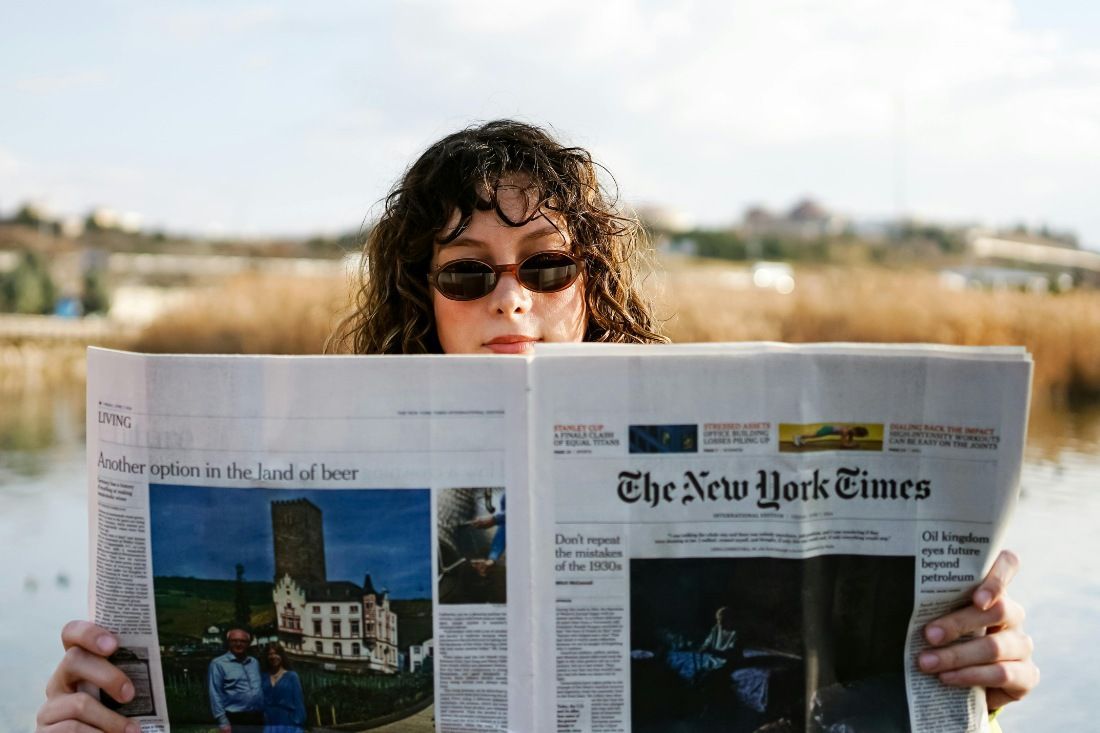 A person with wavy dark hair and sunglasses holds an open New York Times newspaper in front of their face outdoors.