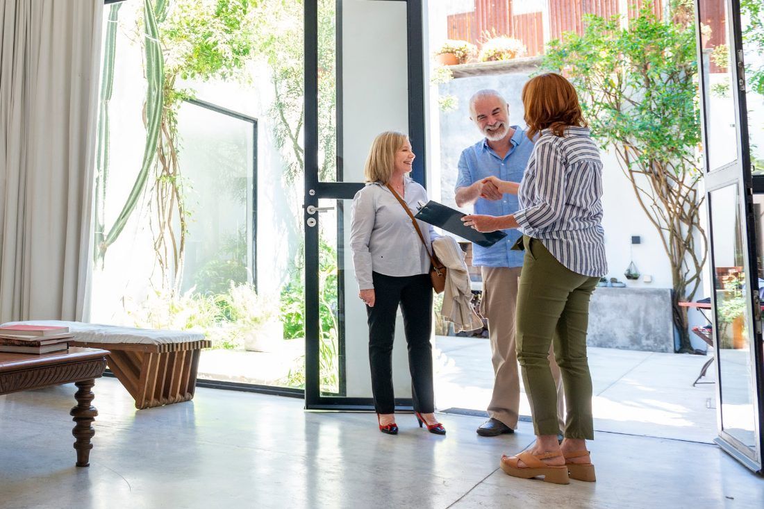 Three people stand in a sunlit doorway, talking and holding a folder, inside a home with an open-air courtyard.