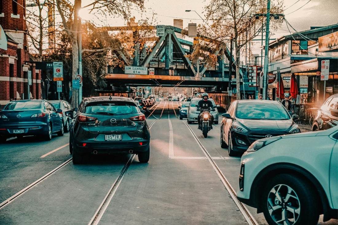 Cars and a motorcycle travel down a city street under a large, arched metal railway bridge at golden hour.