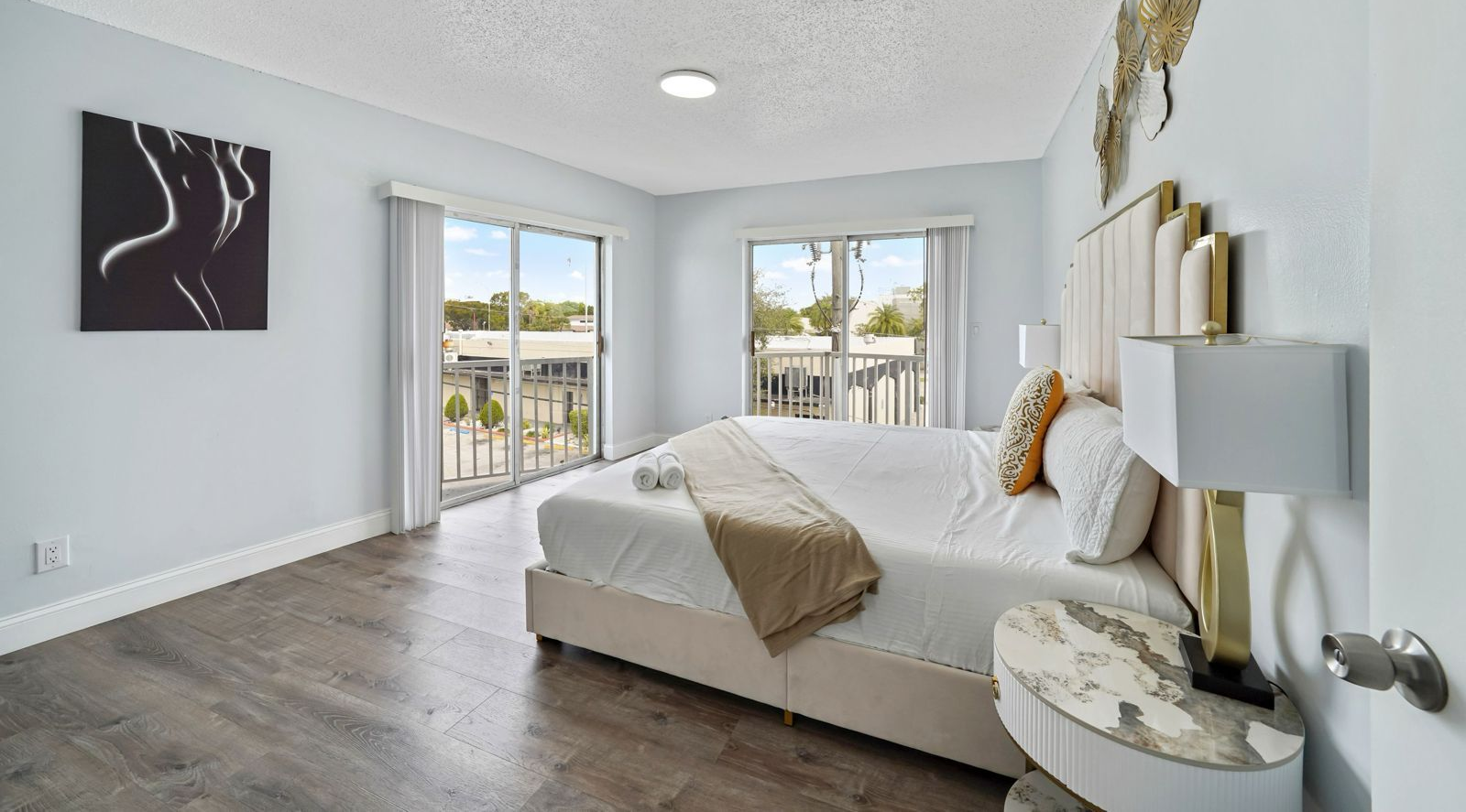 A bright bedroom with a bed featuring a tall beige headboard, dark wood-look floors, and sliding glass doors to a balcony.