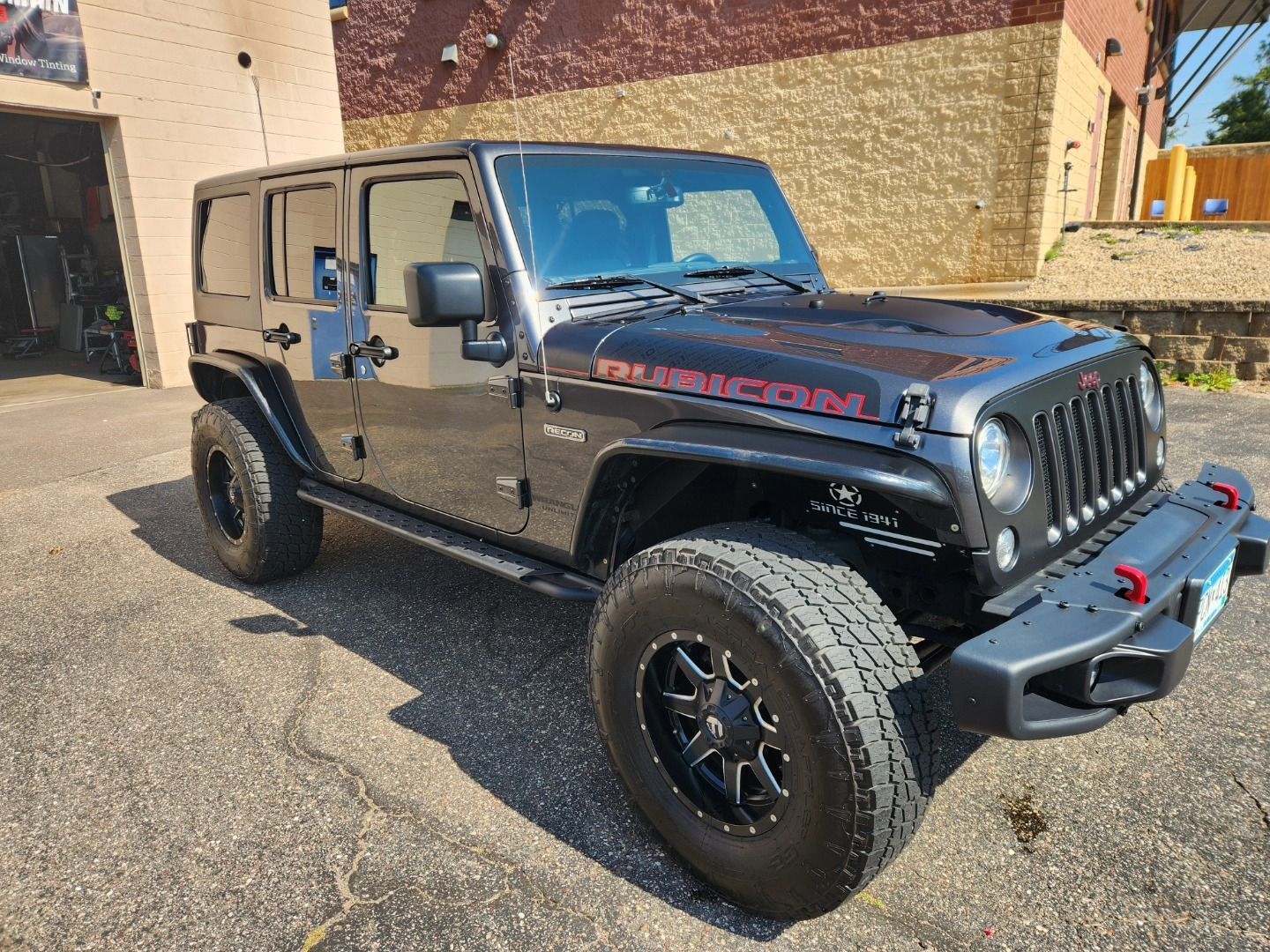Black Jeep Wrangler parked on pavement beside a building, angled front view in sunlight
