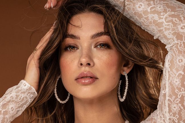 Donna con orecchini a cerchio, mani nei capelli, indossa un top di pizzo bianco, rivolta in avanti.