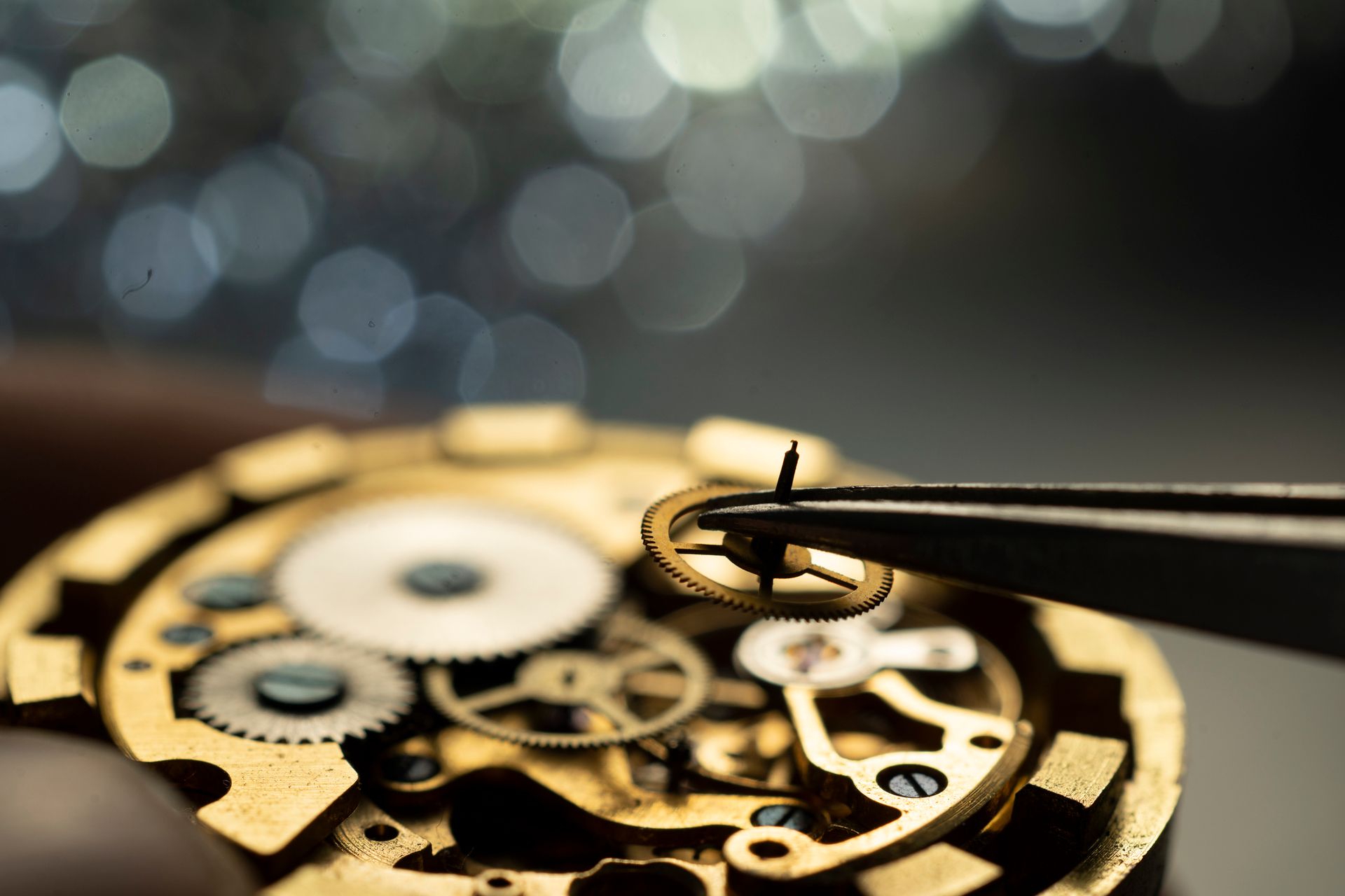 Primo piano degli ingranaggi di un orologio, una mano che tiene un piccolo anello con una pinzetta, sullo sfondo di luci sfocate.