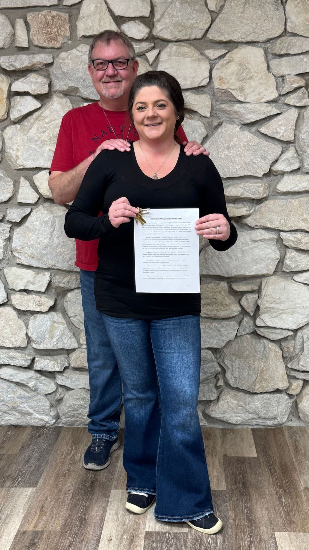 Two people standing indoors against a stone wall, holding a certificate and smiling.