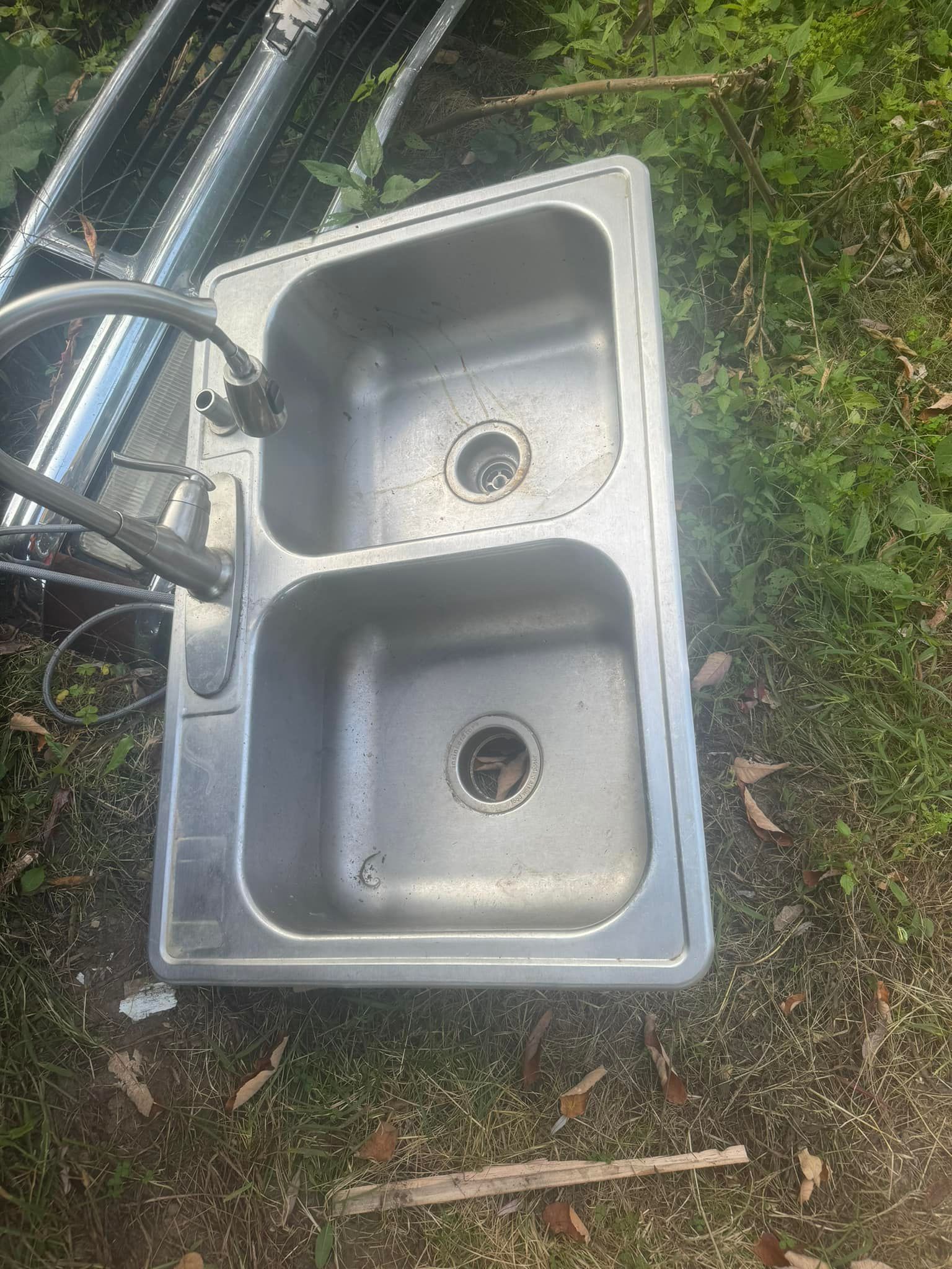 Outdoor stainless steel double-basin sink lying on grass beside hoses and a stick